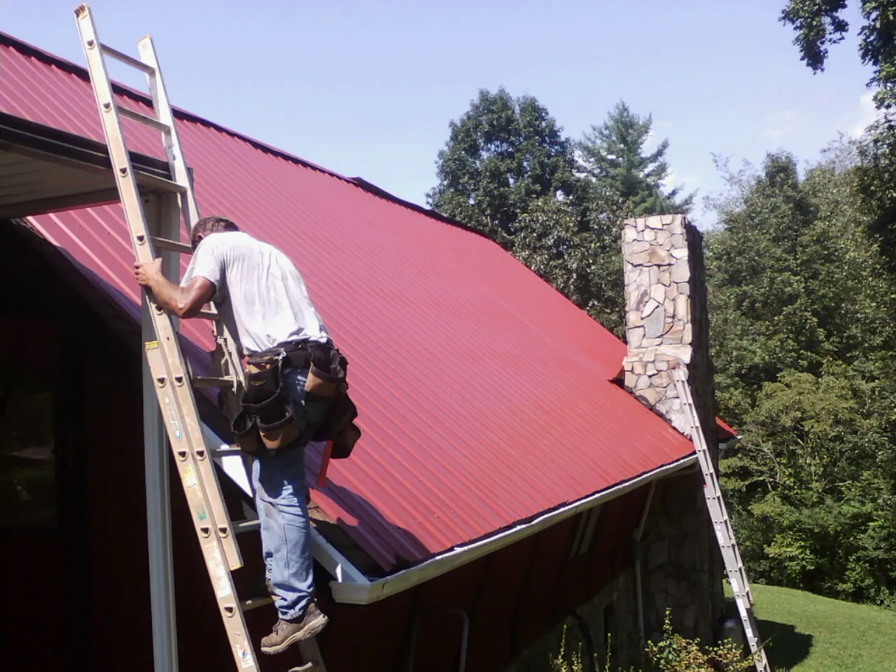 Roofer on a ladder beside a red metal roof and stone chimney — Soffit & Fascia Repair by Roofing Brothers in Higganum